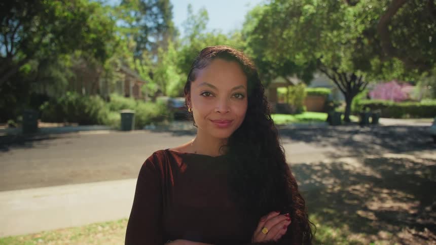 A confident woman with long curly hair crosses her arms and smiles at the camera while standing in a sunny suburban neighborhood