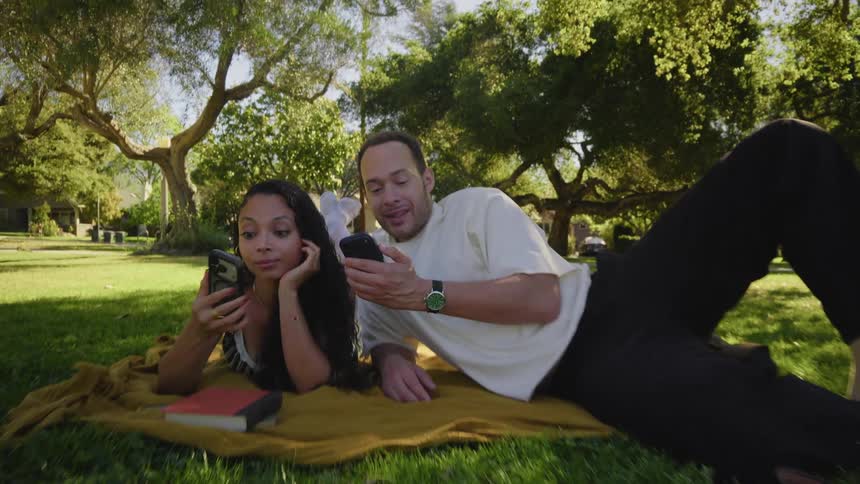 A happy couple lies on a blanket under the trees in a park, smiling while looking at their smartphones together