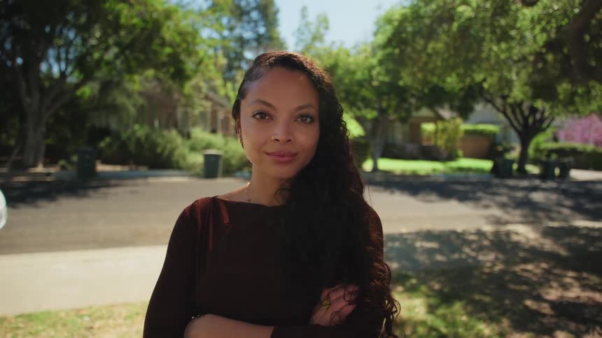 A confident young woman with long curly hair stands on a quiet suburban street and looks directly into the camera