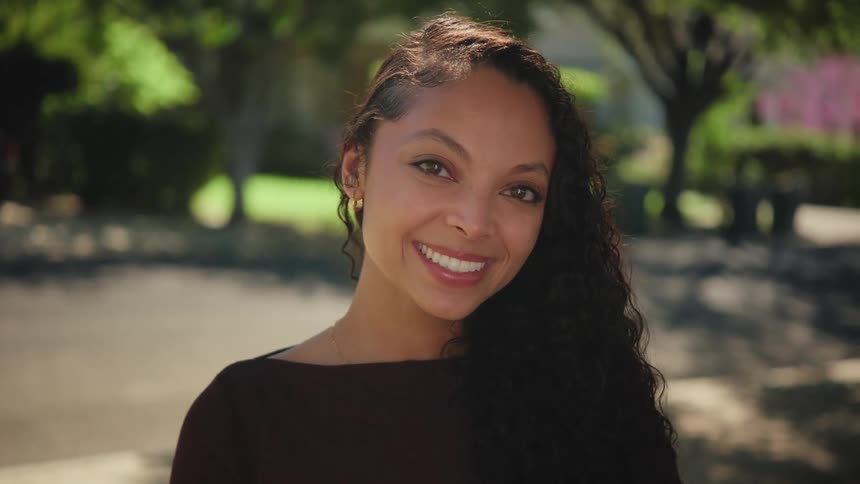 A radiant young woman with long dark hair looks at the camera and breaks into a joyful smile outdoors in slow motion