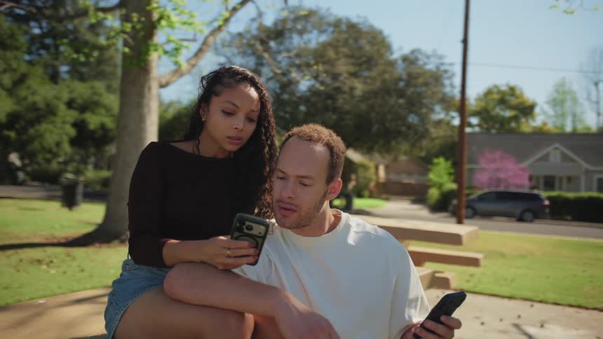 A young couple sits closely together on a park bench, both focused on their phones as they scroll through content on a bright sunny day