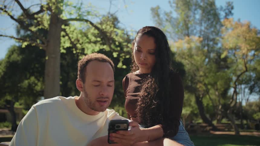 A man shows a woman something on his phone as they relax together in a park on a sunny day