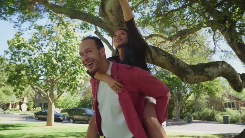 A joyful man gives a woman a piggyback ride under a large tree in a sunny suburban park