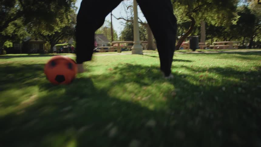 A low-angle point-of-view shot follows a person's bare feet as they dribble a soccer ball across a grassy park on a sunny day