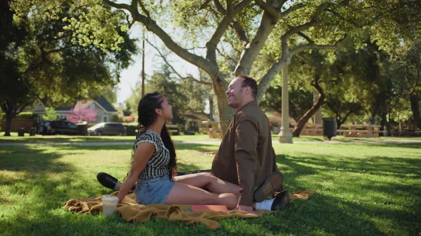 An affectionate couple shares a laugh while sitting on a blanket during a sunny day date in a beautiful park