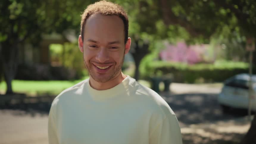 A charming young man smiles and laughs warmly at the camera on a sunny suburban street