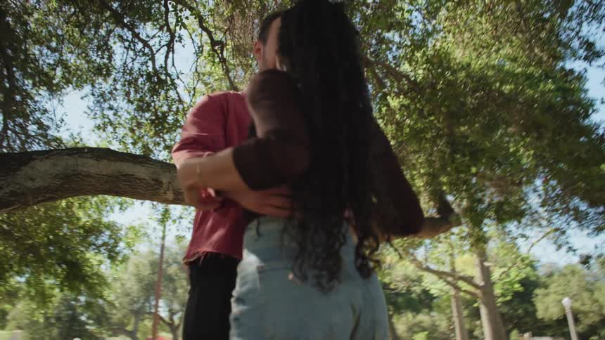 A loving couple slow dances and spins together under a large, sun-drenched tree in a beautiful park