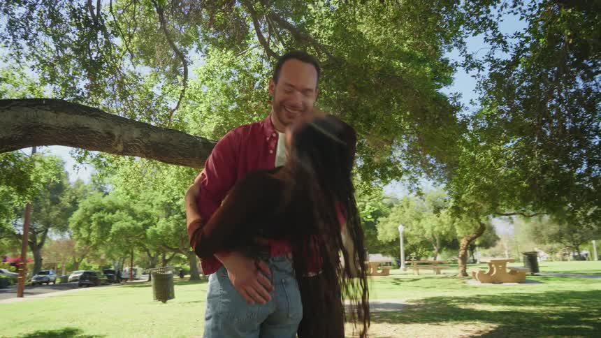 A happy couple dances together in slow motion under a large tree in a sunlit park