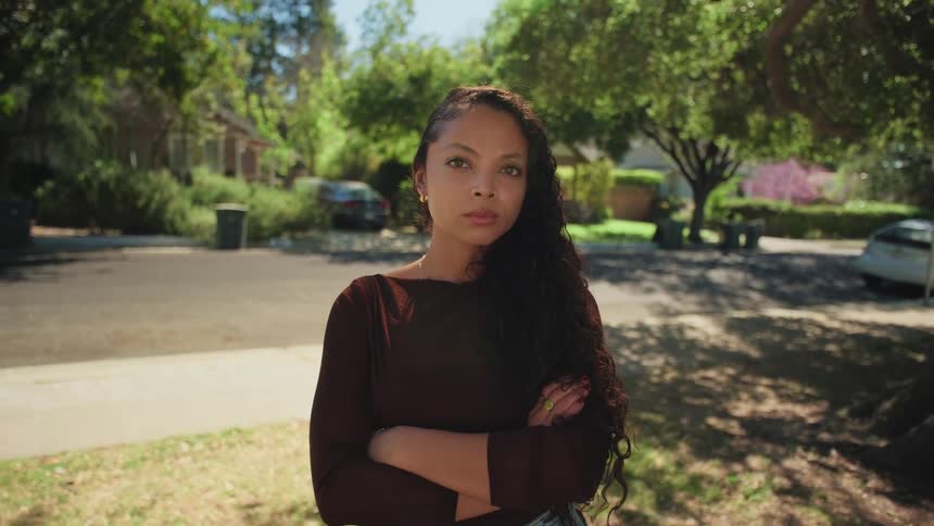 A confident and serious young woman with long curly hair crosses her arms and stares at the camera in a sunny suburban neighborhood