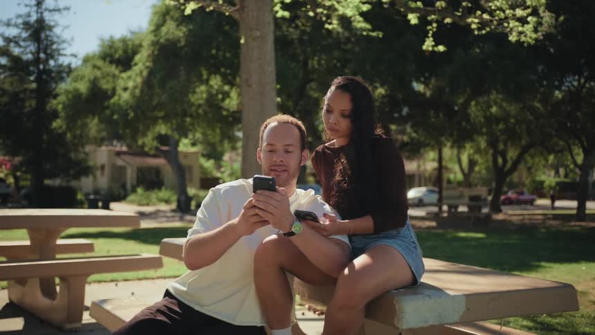 A couple sits closely together on a picnic table at a park, talking and looking at a smartphone screen