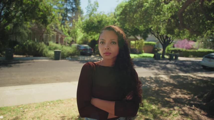 A serious young woman stands on a sunny suburban street, crosses her arms, and looks directly into the camera