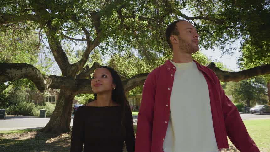 A happy, loving couple smiles and looks at each other while standing under a large tree in a park on a sunny day