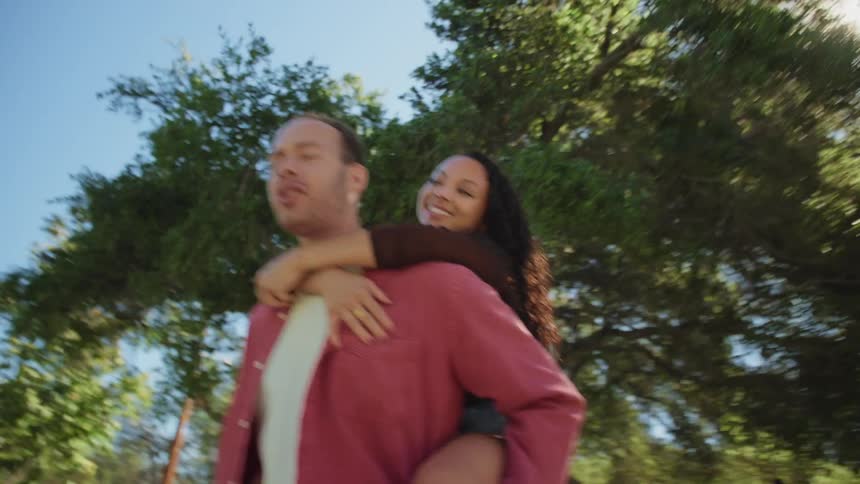 A happy young man gives his partner a piggyback ride as they walk down a sunny, tree-lined street