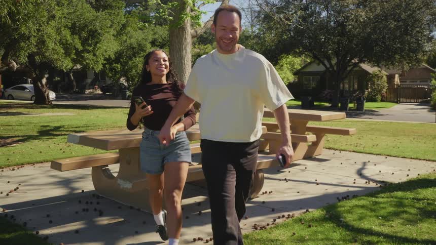 In slow motion, a smiling couple gets up from a picnic table and walks away hand-in-hand through a sunlit park