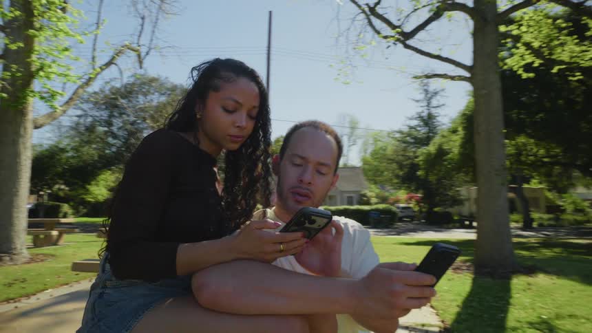 A young couple sits together on a picnic table in a park, both absorbed in their smartphones on a sunny afternoon