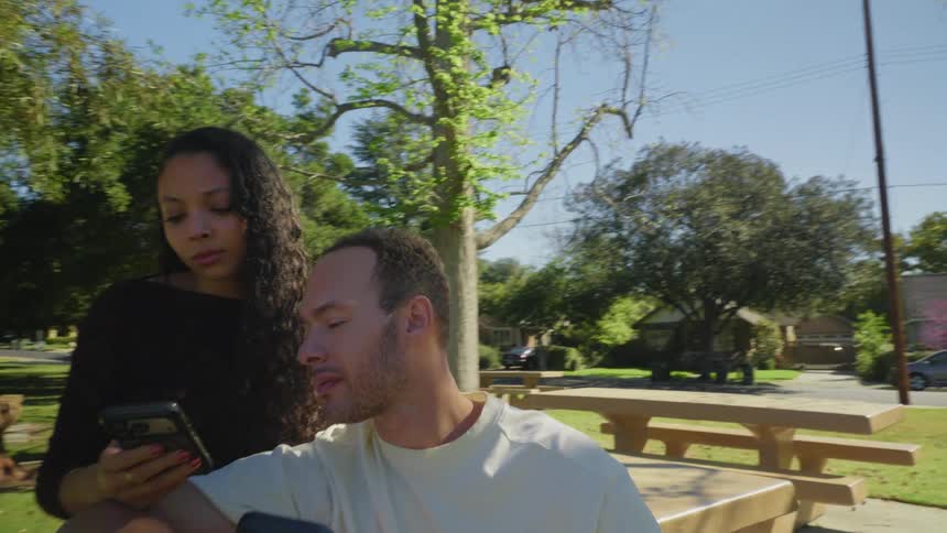 A close-up shot pulls back to reveal a young couple sitting on a park bench, sharing a moment while looking at a smartphone screen together on a sunny day