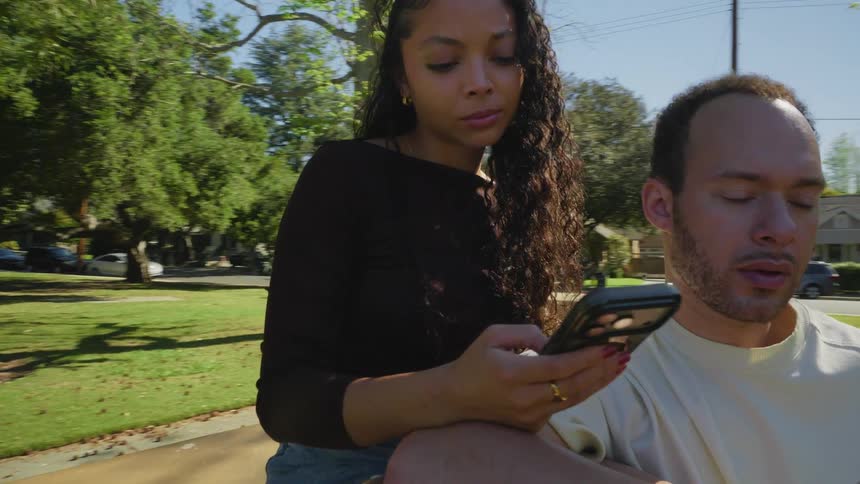 A modern couple sits together at a picnic table in a park, both silently absorbed by their cell phones