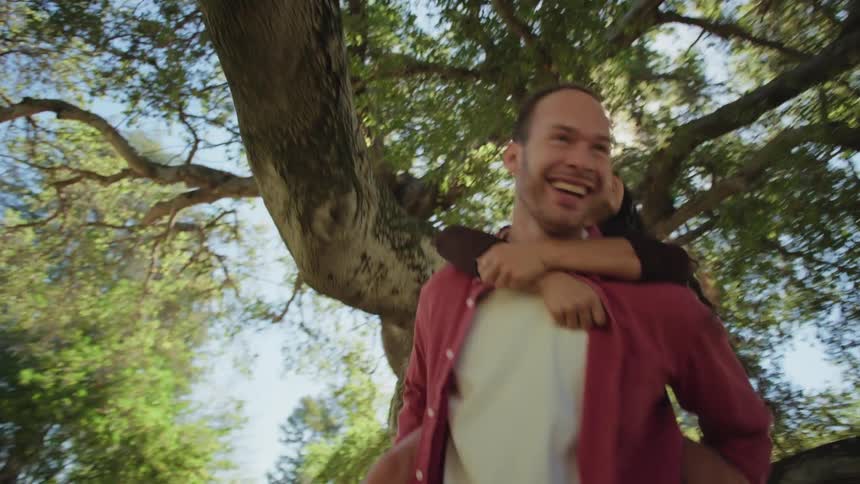 A playful man gives his girlfriend a piggyback ride as they laugh and spin around together under a large tree on a sunny day