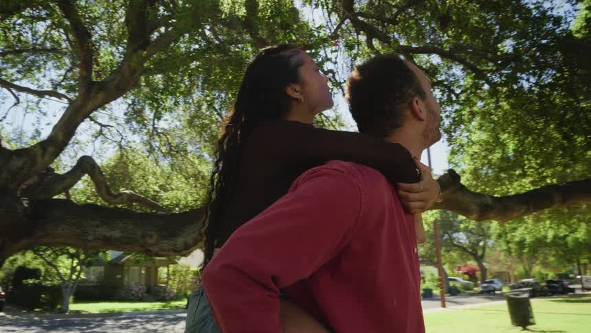 A smiling man gives a woman a piggyback ride as they lovingly gaze up while standing under a large tree in a park