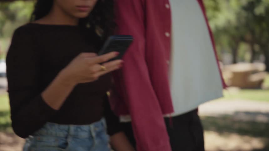 A young woman smiles at her phone while walking hand-in-hand with her partner through a sunny park