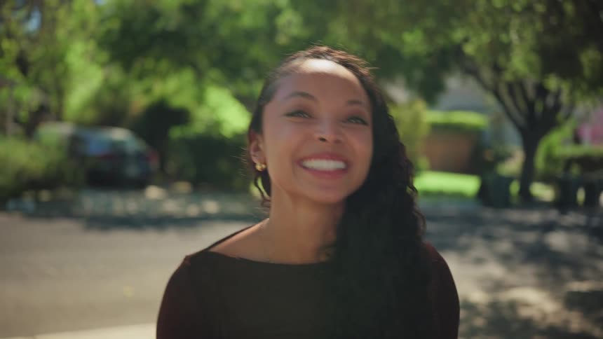 A portrait of a young woman on a sunny suburban street whose somber expression transforms into a joyful, radiant smile
