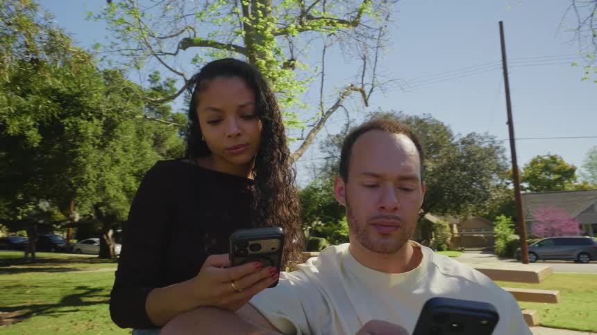 A young couple sits together on a park bench, each engrossed in their own smartphone on a sunny day as the camera slowly pulls back
