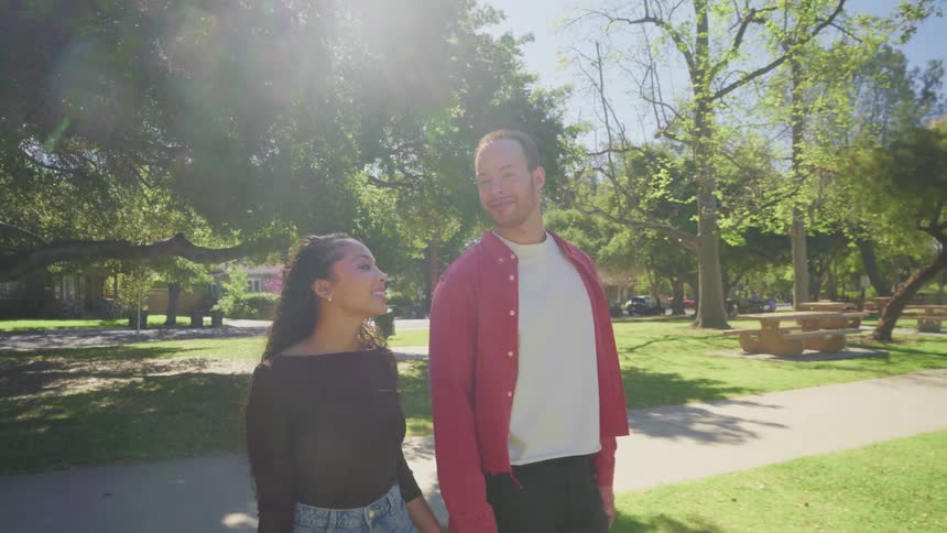 A romantic young couple holds hands and smiles lovingly while walking together through a sun-drenched park in slow motion