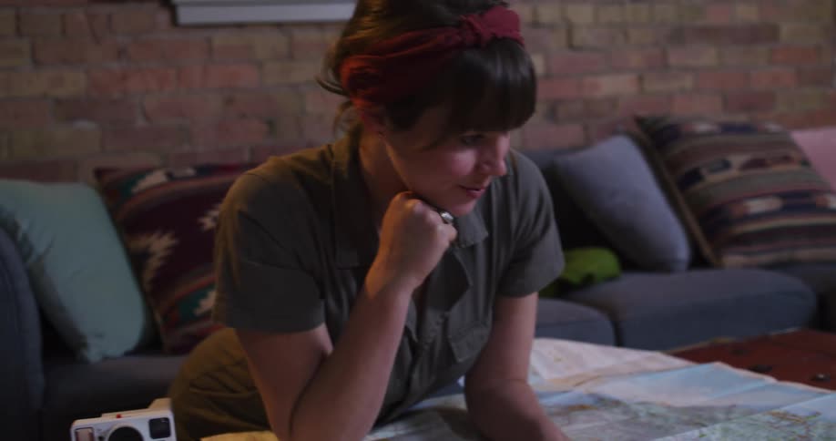 A young woman happily traces a route on a large paper map while planning her next adventure trip in her cozy living room