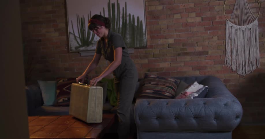 A woman in a retro jumpsuit arrives at her loft apartment, opens a vintage suitcase, and smiles as she walks away