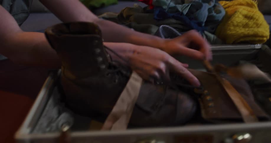 A woman prepares for her next adventure by carefully packing her rugged boots and clothes into a vintage suitcase