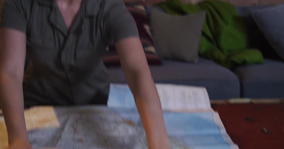 An adventurous woman in a khaki jumpsuit dramatically unfolds a large road map onto a coffee table to plan her next journey