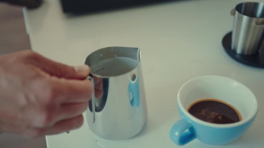 A close-up of a barista swirling steamed milk in a metal pitcher next to a cup of fresh espresso