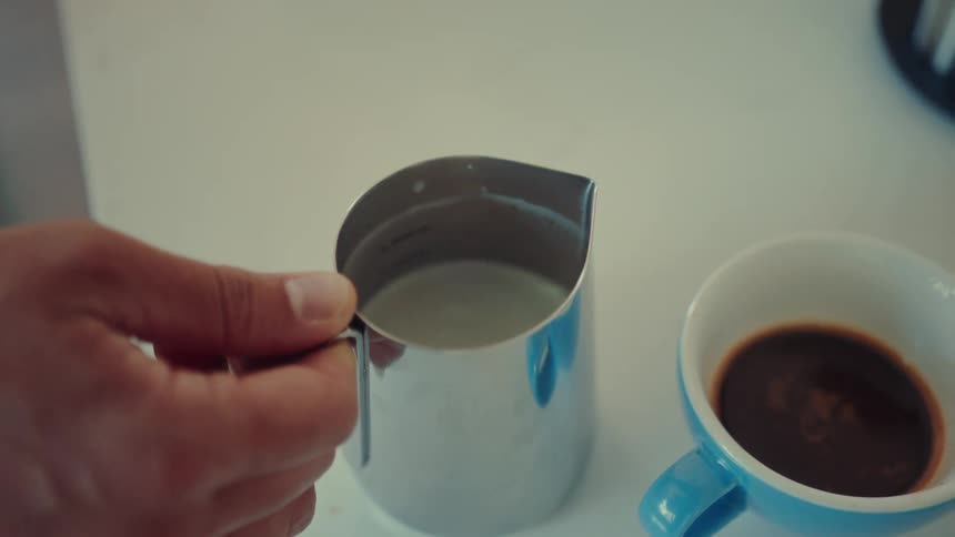 A skilled barista's hand swirls hot, foamy milk in a stainless steel pitcher in slow motion, preparing to create latte art