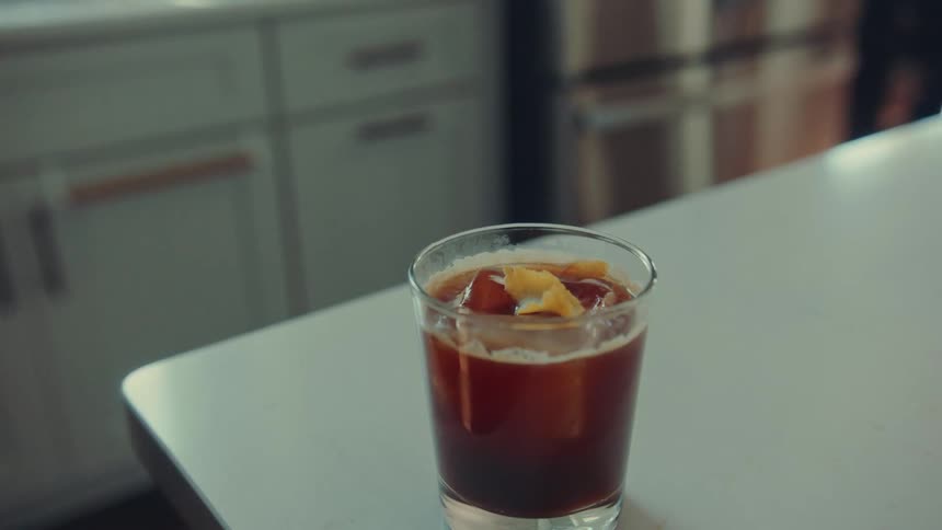 A hand carefully garnishes a glass of iced coffee with a fresh orange peel in slow motion