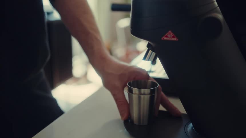 A person grinds fresh coffee beans into a metal dosing cup with an electric grinder in a slow-motion close-up