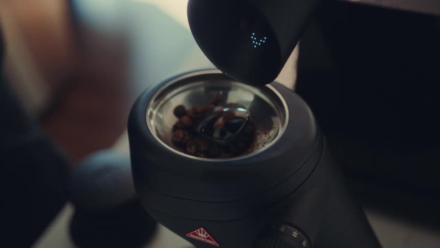 A close-up slow-motion shot of a barista pouring whole roasted coffee beans into an electric grinder