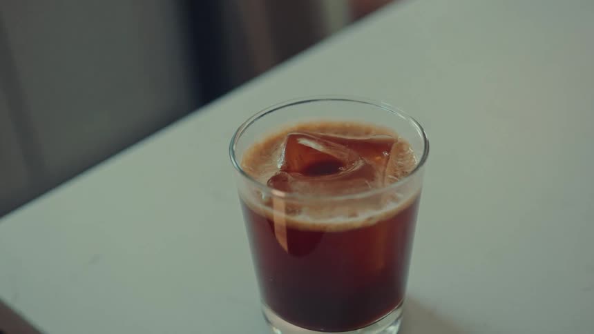 A cinematic close-up of a glass of iced coffee settling on a bright tabletop before the camera goes out of focus