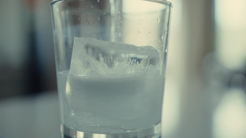 A time-lapse shows a large, clear ice cube slowly melting in a glass, with condensation forming on the outside