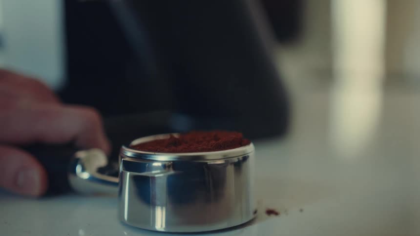 A barista prepares espresso by using a WDT tool to distribute fresh grounds in a portafilter and then tamping them down, all in cinematic slow motion
