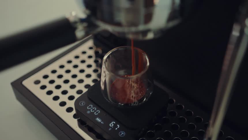 A close-up shot captures fresh, dark espresso pouring from a machine into a glass cup on a digital scale