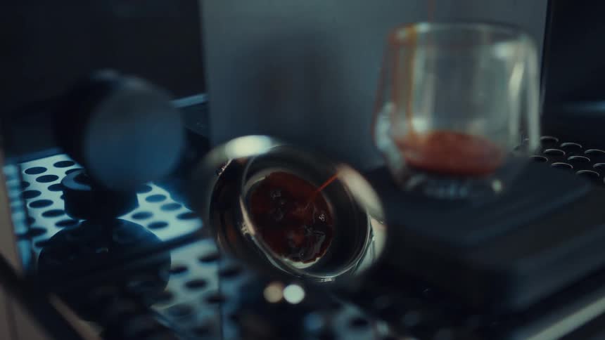 A dramatic close-up of dark espresso slowly dripping from a portafilter and into a glass cup