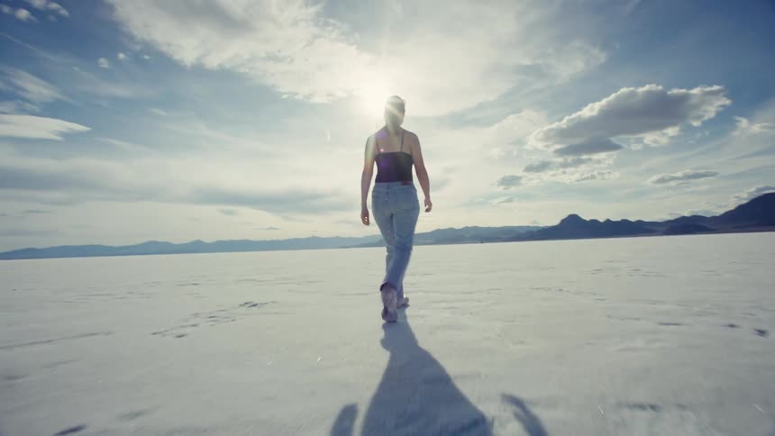 A slow-motion tracking shot follows a woman walking away across the expansive Bonneville Salt Flats under a bright, sunny sky