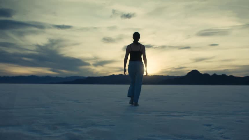 A lone woman walks across a vast, empty salt flat at sunset towards distant mountains