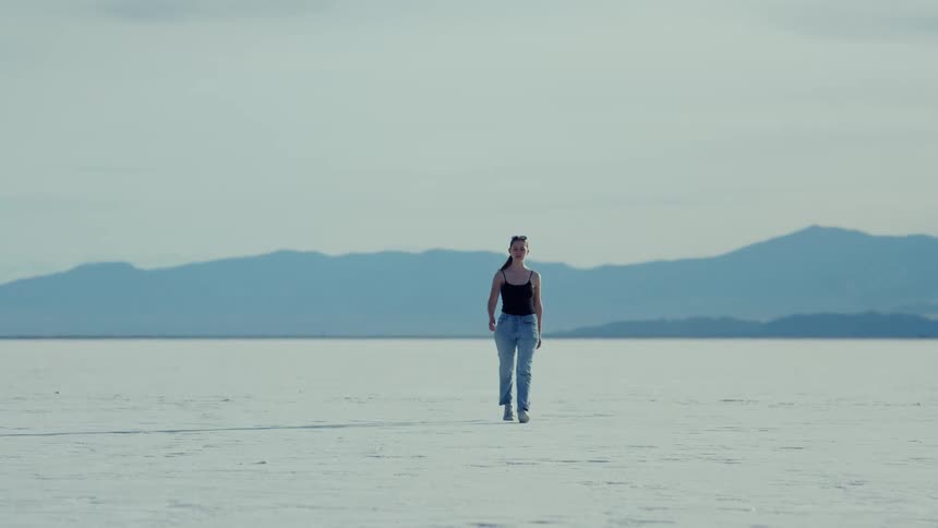 A woman slowly comes into focus as she walks across a vast, white salt flat with mountains in the background