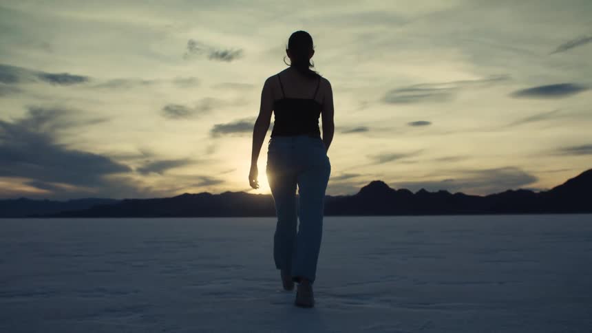 Slow motion shot from behind of a woman's silhouette as she walks across a vast, empty desert landscape towards the setting sun