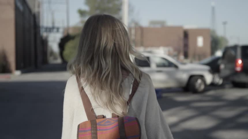 A blond woman in a brown sweater carrying a colorful backpack at walks through a parking lot