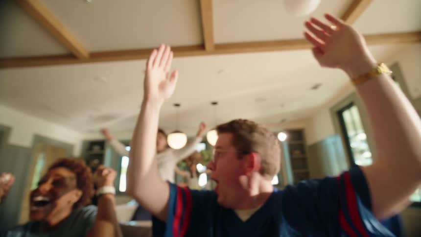 Excited sports fans leap from their couch and cheer in slow motion as popcorn flies through the air during a game-winning play