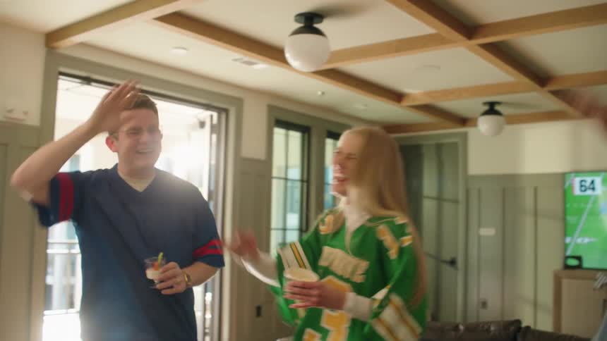 A group of diverse friends in football jerseys cheer and high-five in slow motion while watching a game at a party