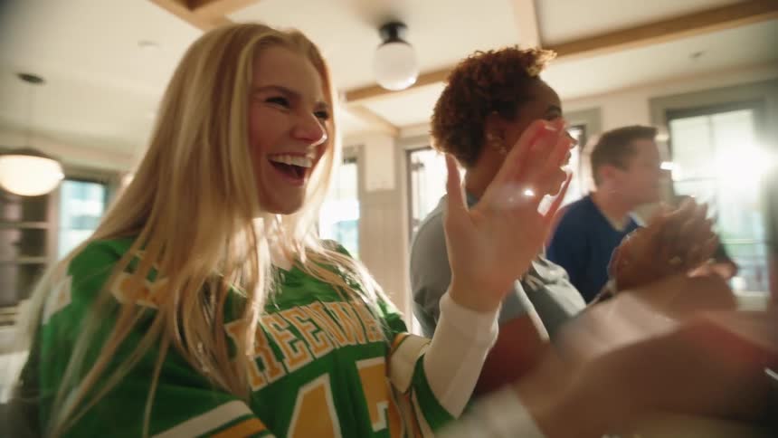 An excited group of diverse friends in football jerseys clap and cheer in slow motion while watching a sports game at a sunlit party
