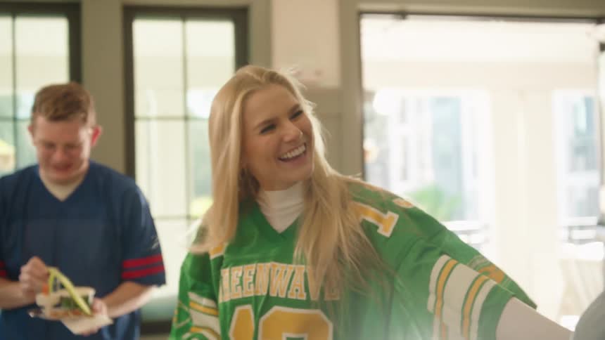 Friends in football jerseys get food and laugh together at a gameday watch party in slow motion